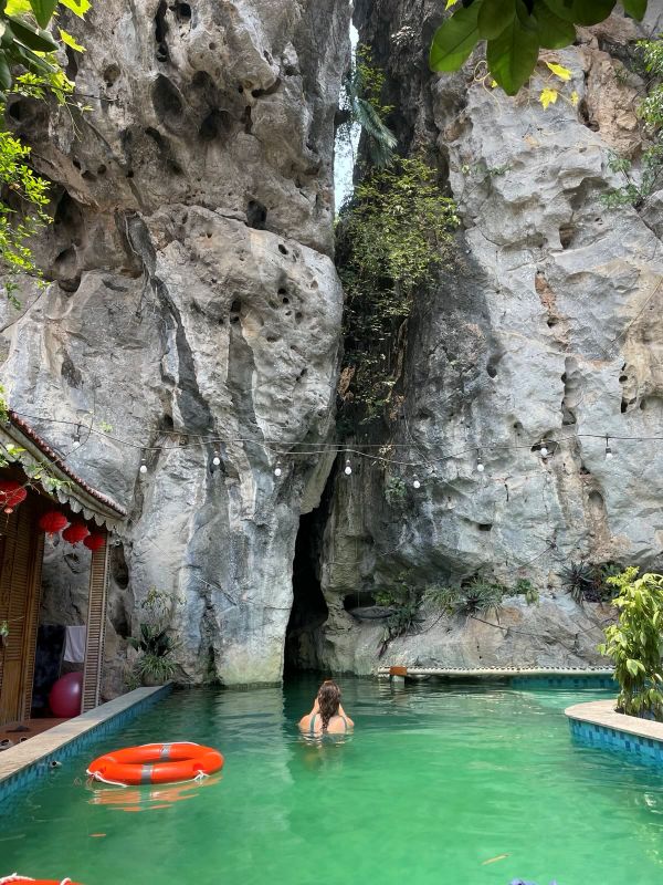 "Ein Swimming Pool mit grünem Wasser, in dem ein oranger Schwimmreif schwimmt. Daneben ist Sophia, wie sie der Kamera den Rücken zugewandt hat und ein Foto von dem großen Spalt des Felsens macht, in den der Pool hineinragt."