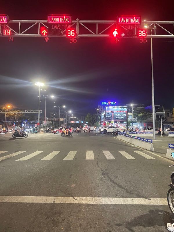 "An einer Kreuzung queren einige Mopeds die Straße. Im oberen Bereich befindet sich eine Ampelanlage mit drei roten Ampeln und der Sekundenanzeige die "36" zeigt."