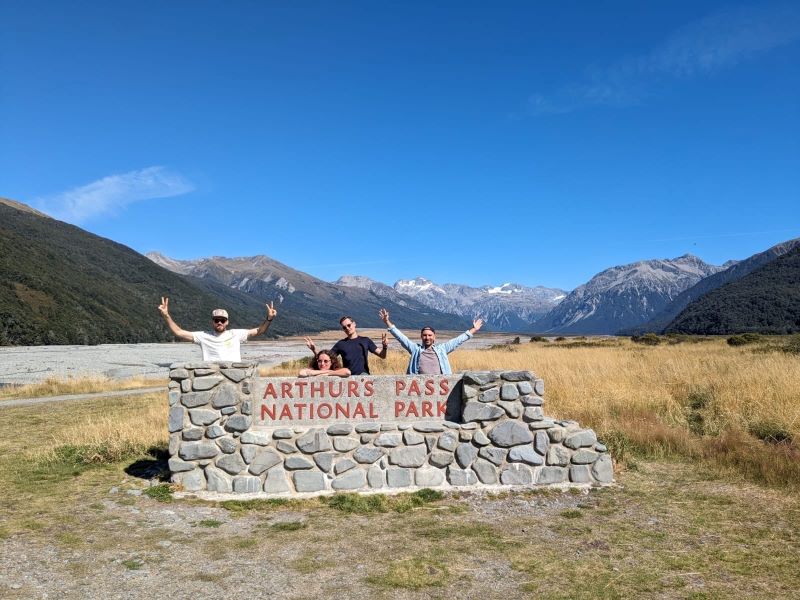 "Wir posieren als Gruppe vor dem Arthur's Pass National Park Schild mit dem Bergen im Hintergrund"