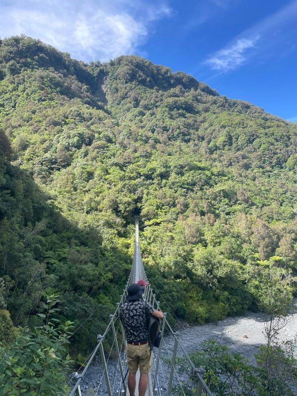 "Simon und Sophia auf einer Hängebrücke. Hinter ihnen ein großer Wald unter über ihnen ein blauer HImmel."