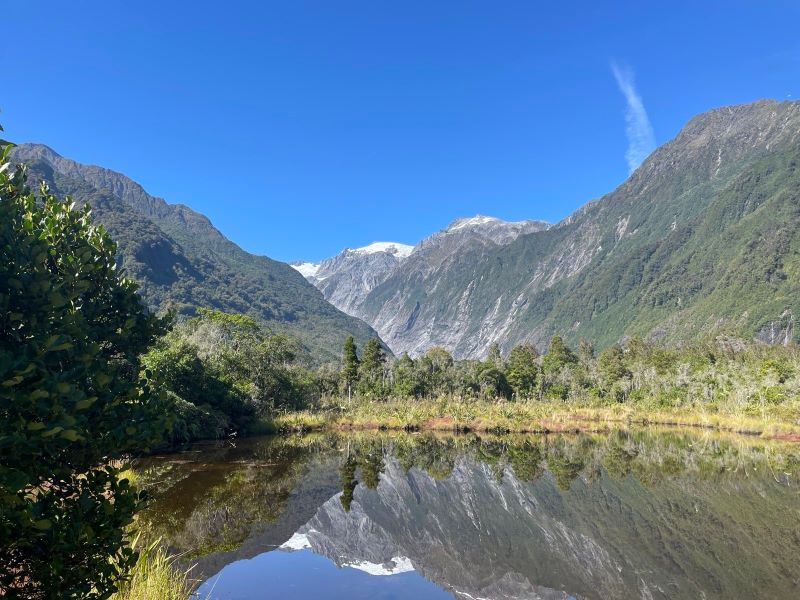 "Bergige Landschaft bei blauem Himmel mit schneebedeckten Gipfeln und einer bewaldeten Landschaft, die sich in der Reflektion eines stillen Sees spiegelt."