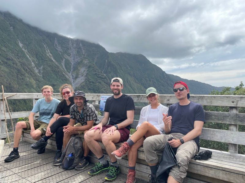 "Eine Gruppe von sechs Leuten, wie sie auf einer Aussichtsplattform aus Holz posieren. Dahinter eine bewaldete Berglandschaft und bewölkter Himmel."