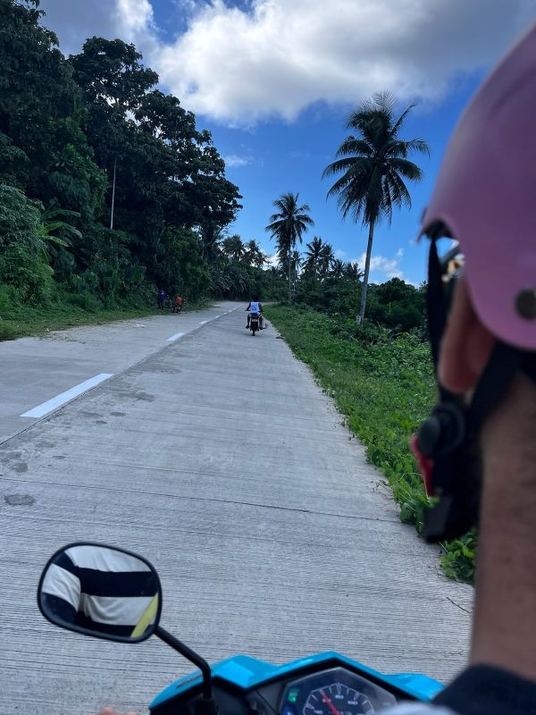"Blick über meine Schulter, als ich Moped fahre. Man erkennt meinen Helm sowie den Rückspiegel und die Konsole des Mopeds. Ich fahre auf einer Straße David hinterher. Am Straßenrand ist grüne Vegetation."