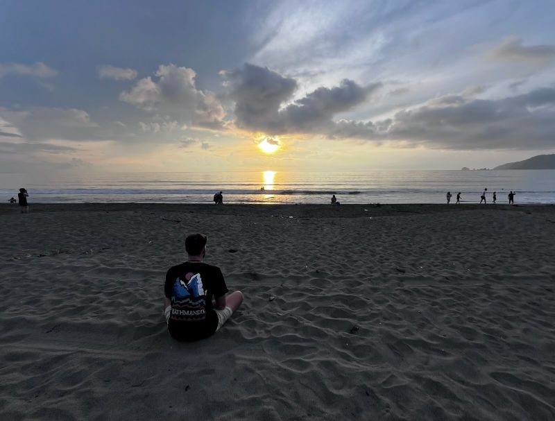 "Der junge Mann sitzt im Schneidersitz am weichen Sandboden. Er trägt ein schwarzes T-Shirt, auf dem eine Illustration von zwei Bergen zu sehen ist, zwischen denen die Sonne aufgeht. Im seichten Wasser spazieren eine handvoll Menschen entlang. Dahinter, über dem Meer, geht die eigentliche Sonne auf."