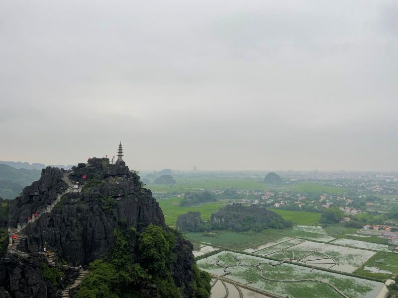 "Ein Berg, auf den sich Stufen aus Stein nach oben winden. An der Spitze befindet sich ein kleiner Turm. Unter dem Berg sind mehrere Reisfelder, über die Wege verlaufen."
