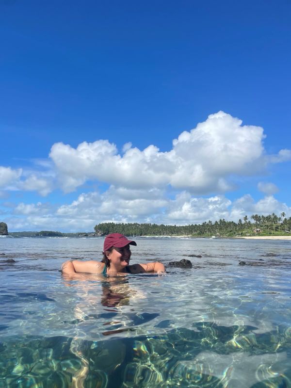 "Sophia stützt sich mit den Armen am Rand des Felsbeckens ab und lächelt. Sie ist bis zum Hals im Wasser und hinter ihr erkennt man das Meer."