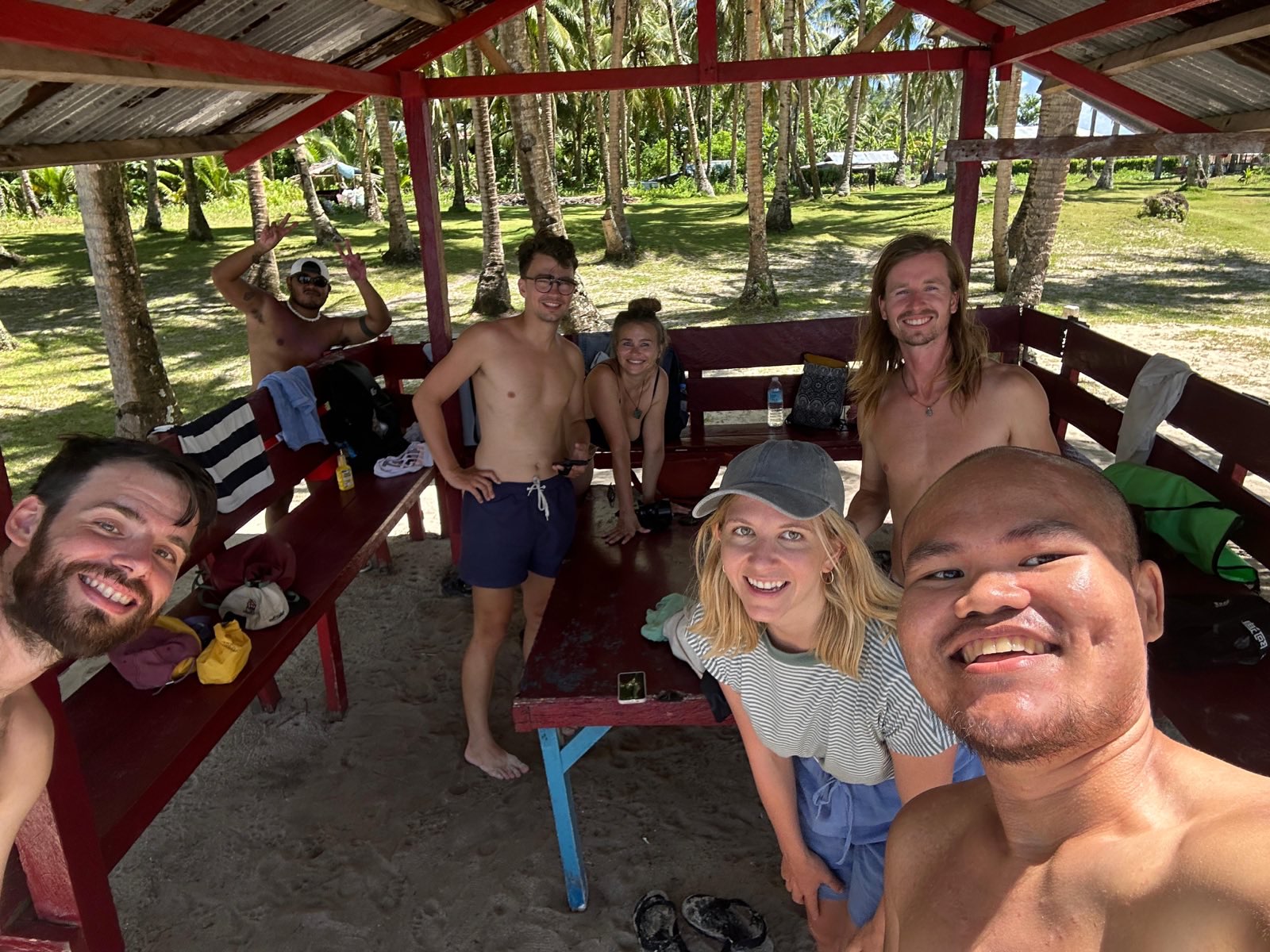 "Unsere Gruppe unter dem Strandhaus. Ich lächle in die Kamera, Miguel hebt seine Hände zum Peace-Zeichen. Philippe, Jennifer, Henrik, die andere Sophia und Wesson lächeln. Einige von uns sind in Badekleidung."