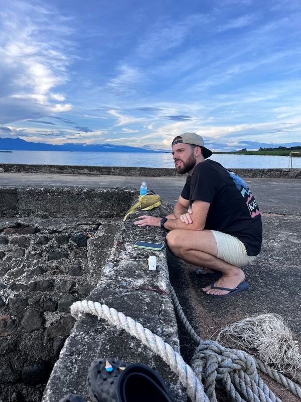 "Ich hocke am Port und blicke nachdenklich aufs Meer. Ich trage ein schwarzes T-Shirt, eine kurze beige Hose und mein beiges Basecap."