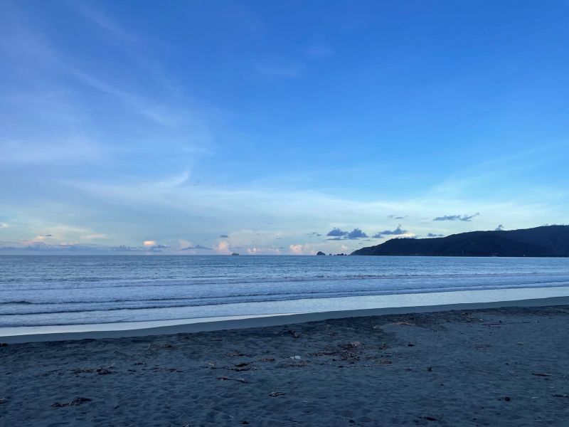 "Blick vom Strand aus aufs Meer. Neben den Bergen und den Felsen ist ein Schiff am Horizont zu erkennen."
