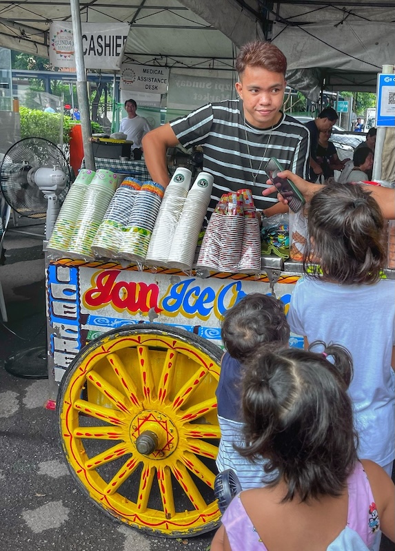 "Ein junger Filipino an einem Getränkestand. Er trägt ein gestreiftes T-Shirt und eine Halskette. Er hat einen schmunzelnden Blick aufgesetzt."