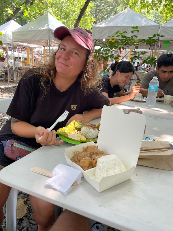 "Sophia lächelt am Esstisch. Vor ihr steht ein Silog auf grünem Teller und daneben mein Sizzling Tofu in einer Papierbox."
