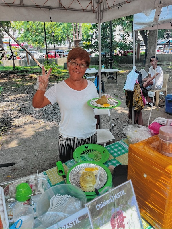 "Die Frau vom Essensstand hält eine Hand zum Victory-Zeichen in die Luft und hält in der anderen einen grünen Teller mit Silog. Sie lächelt freundlich."