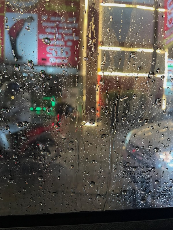 "Hinter einer nassen Fensterscheibe erkennt man ein Moped und ein Auto auf der Straße. Ein Schild zeigt die Illustration eines sich verbeugenden Mannes in rotem Anzug."
