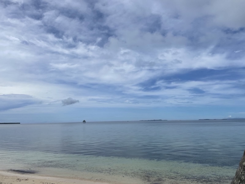 "Das blaue Meer mit wolkenverhangenen Himmel. In der Ferne erkennt man eine kleine Holzhütte am Wasser."