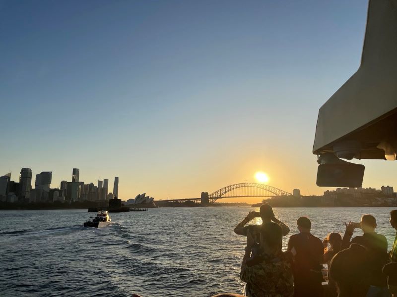 "Die Sonne geht unter über der Harbour Bridge. Im Vordergrund sind Menschen auf der Fähre zu sehen wie sie Photos machen."