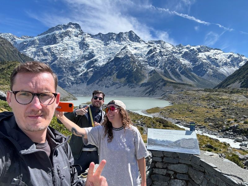 "Manu, Sophia und ich lächeln. Während Manu zwei Finger zur Pose in die Kamera hält, machen Sophia und ich dahinter einen Selfie. Im Hintergrund sieht man eine grüne Landschaft, den Gletschersee und den schneebedeckten Mt. Cook."
