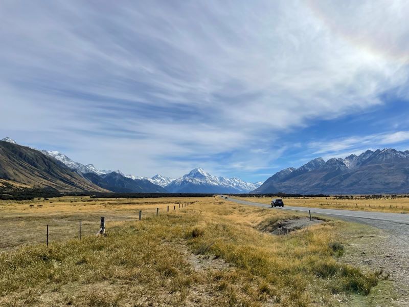 "Eine Straße, umgeben von braun-gelben Büschen führt zum Mt. Cook. Die Berglandschaft ist hügelig und die Berge sind schneebedeckt. Der Himmel ist leicht bewölkt."