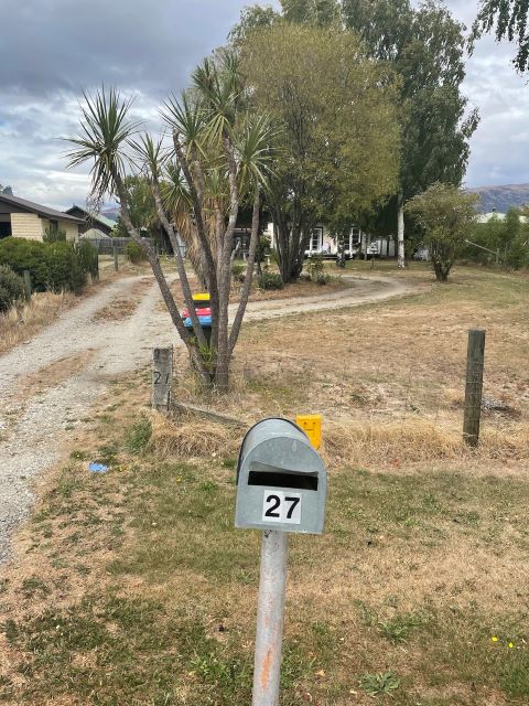 "Eine Einfahrt führt an einem Postkasten vorbei in ein typisches Grundstück in Albert Town. Links neben der Einfahrt sind einige grünen Büsche, rechts daneben ein Baum, um den die Spur herumführt. Der Garten hat braunes Gras und einige Palmenbäume."