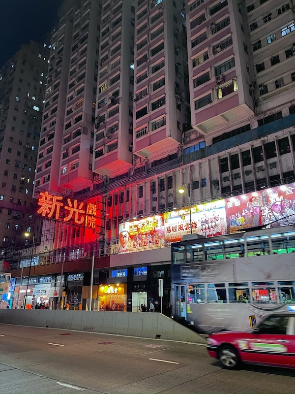 "Eine Straße in North Point bei Nacht. Ein rotes Taxi fährt an der Straße. Darüber ist ein Hochhaus mit Neon-Beleuchtung."