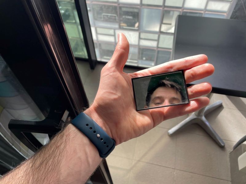 In meiner Hand halte ich einen kleinen Handspiegel mit schwarzem Rahmen. Im Hintergrund erkennt man einen Balkon mit grauen Fließen und einem Plastiktisch. Am Handgeländt trage ich eine Smartwatch mit blauem Band. In der Spiegelung des Handspiegels erkennt man mein Gesicht.