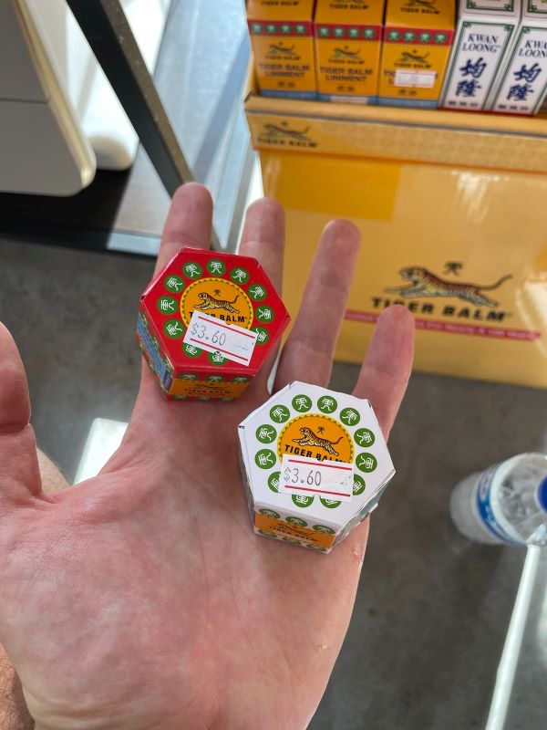 In meiner Hand halte ich zwei kleine Kartons von Tigerbalm, einmal in roter und einmal in weißer Verpackung. Auf beiden ist die Illustration eines Tigers und der Schriftzug "Tiger Balm" zu erkennen. Außerdem ist auf beiden ein Preisschild angebracht: $ 3.60