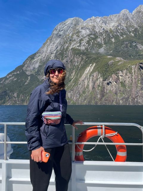 "Sophia steht in blauer Jacke mit zugedeckter Kaputze und Sonnenbrille auf einem Boot. Diagonal um ihre Brust trägt sie eine Bauchtasche in Neonfarben: rot, türkis und lila. Hinter ihr ein blauer See und ein Berg unter einem blauen Himmel."