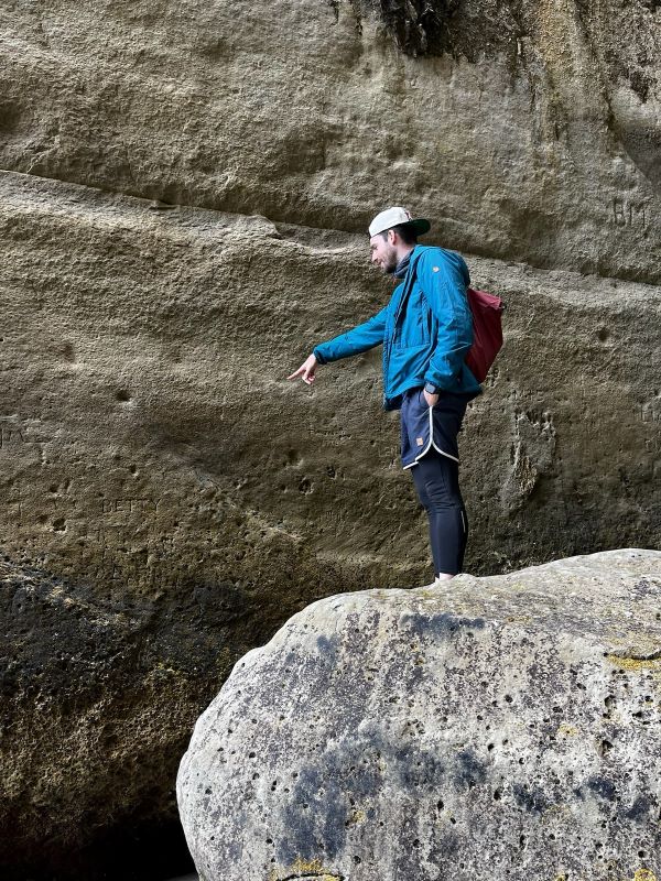 "Ich stehe vor einer Felswand auf einem großen Felsen und zeige mit dem Finger vor mich. Auf dem Rücken trage ich einen burgunderroten Rucksack"