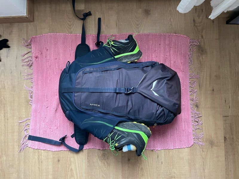 Ein blauer Rucksack liegt mit geschlossenem Reißverschluss auf dem Parkettboten. In den Seitenfächern befindet sich jeweils ein schwarzer Wanderschuh mit grün-gelben Schnürsenkeln.