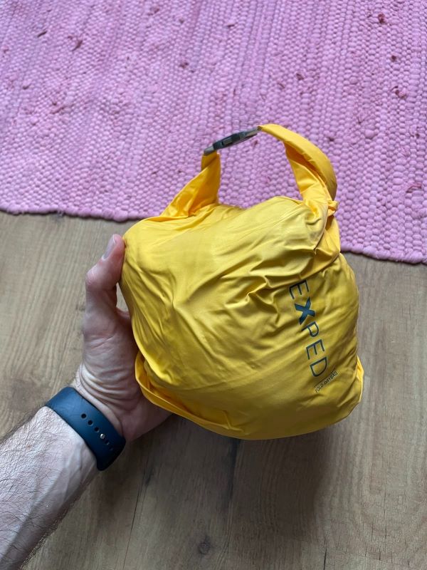 Auf dem Parkettboden halte ich ein gelbes Drybag in der Hand, in dem die Jacke kompakt als Knäuel verdichtet wurde.