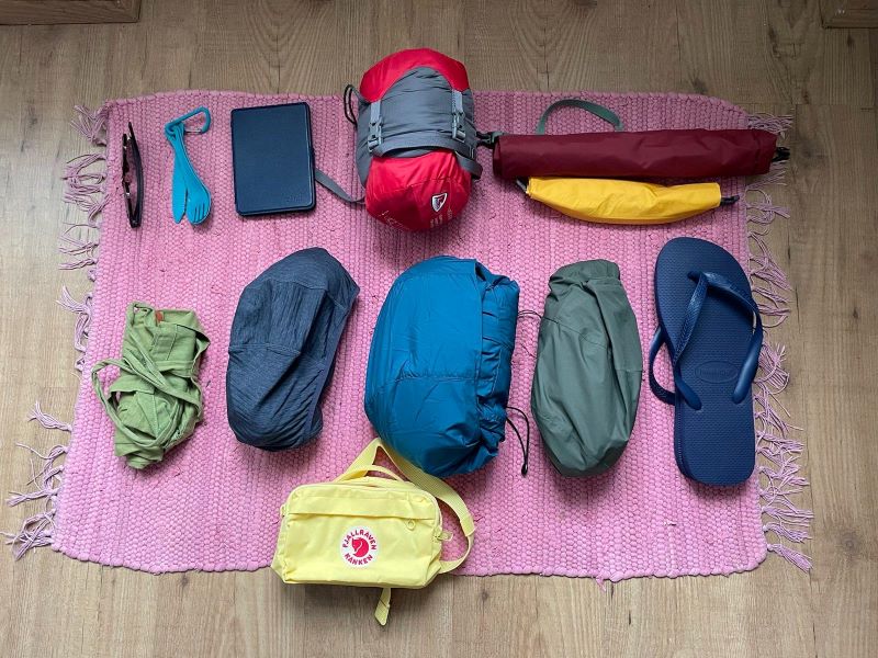 Auf einem roten Stoffteppich liegen folgende Gegenstände: eine Sonnenbrille, türkises Plastikbesteck, ein rot-grauer, zusammengerollter Schlafsack, ein roter, zusammengerollter Rucksack über einem gelben, zusammengerollten Drybag, ein grüner zusammengerollter Jutesack, eine graue zusammengerollte Fleecejacke, eine blaue, zusammengerollte Jacke, eine grüne, zusammengerollte Regenjacke, blaue Flipflops und eine gelbe Bauchtasche.