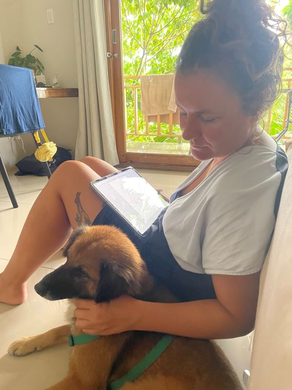 "Sophia sitzt am Fliesenboden der Unterkunft. Am Schoß liegt ihr iPad und mit der linken Hand streichelt sie den braunen Hund "Brownie". "