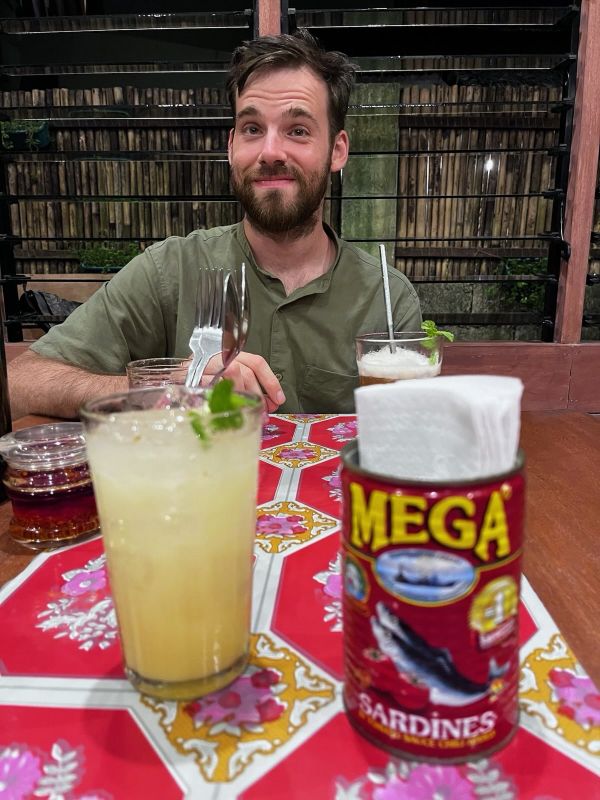 "Ich sitze zu Tisch und lächle verzwickt. Der Tisch hat ein rotes, Mandala-artiges Muster und darauf stehen zwei Getränke, Besteck und eine Sardinendose, mit der Aufschrift "Mega", die als Serviettenhalter dient."