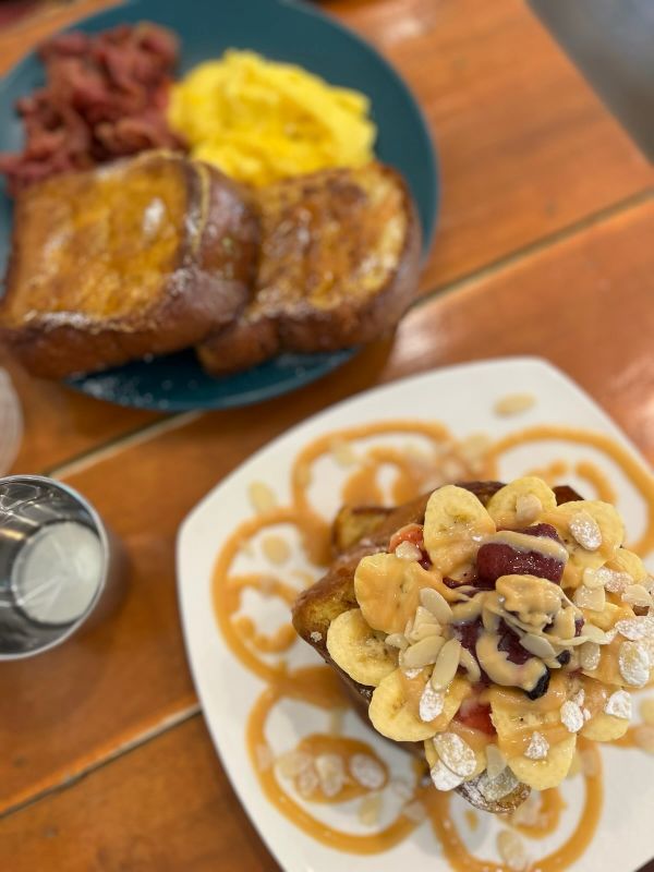 "Dicke Scheiben French Toast, auf einem Teller mit Omlette und Speck, auf dem anderen Teller mit Bananen, Mandeln, Rosinen und Sirup"