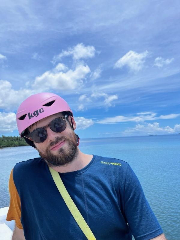 "Porträt-Bild von mir, mit rosa Helm und Sonnenbrille. Hinter mir das blaue Meer und der blaue Himmel mit ein paar Wolken"