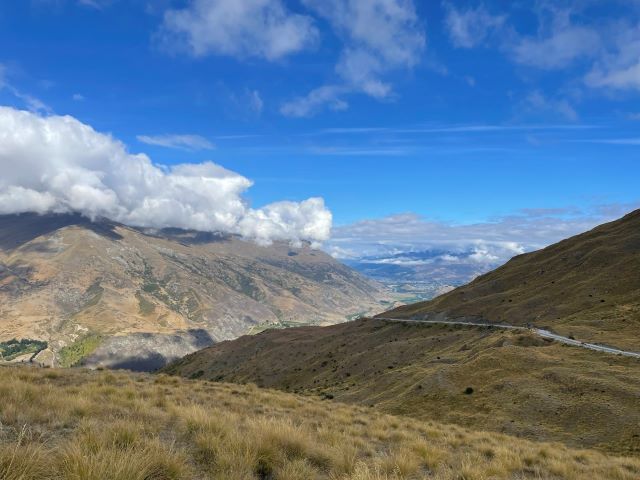 "Blick ins Tal über braune Hügel und dunkelgelbe Büsche. Der Himmel ist blau und zeigt eine Wolkenformation über den braunen Bergen."