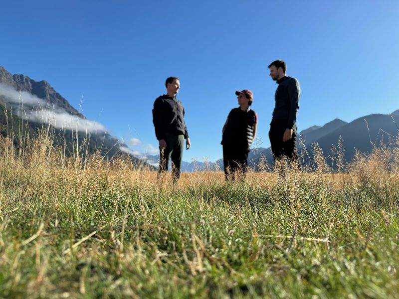 "ine Graslandschaft mit grünen und gelben Wiesen geht in ein Tal hinein. Leicht verschwommen sieht man unsere schwarzen Silouetten im Vordergrund: Manu, Sophia und mich"