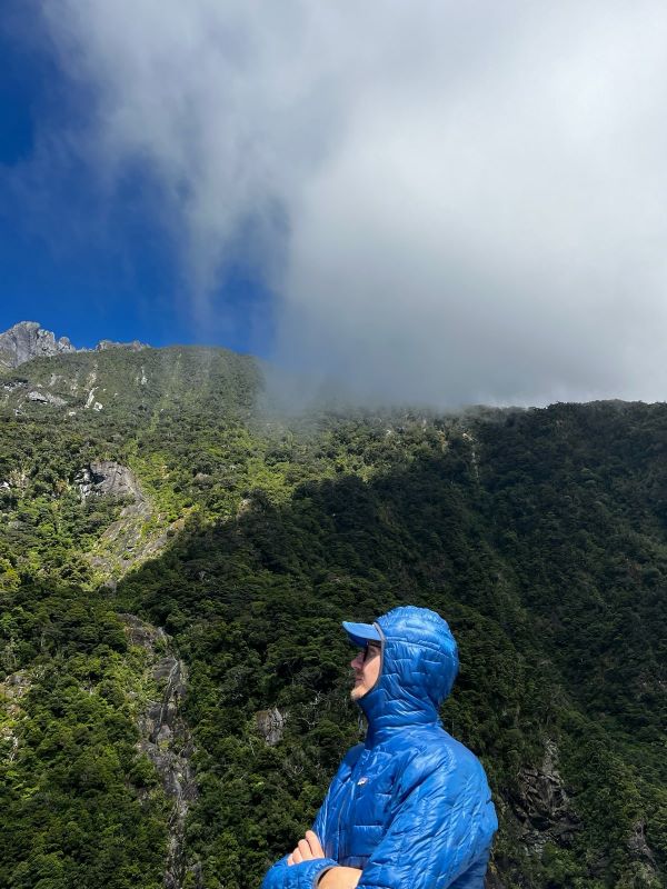 "Manu steht in seiner blauen Jacke auf dem Boot. Er schaut nach links. Im Hintergrund ist eine Berglandschaft und eine weiße Wolke am Himmel zu erkennen."