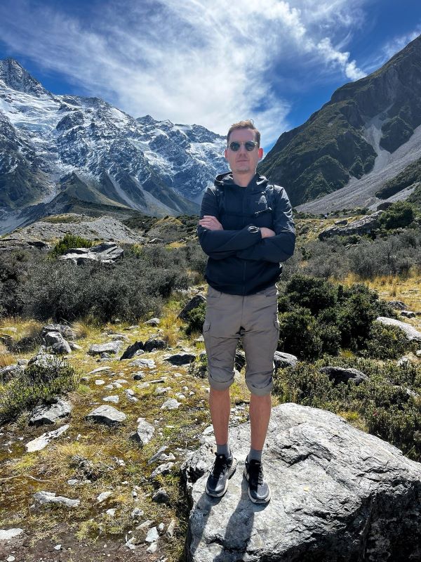 "Einige Wolken über schneebedeckten Bergen. Manu steht im Vordergrund mit verschränkten Armen."