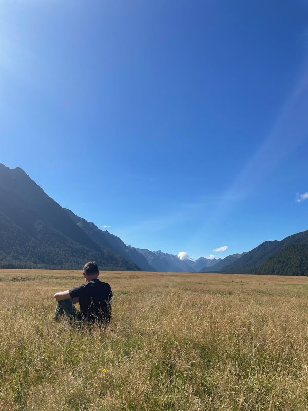 "Eine Graslandschaft mit einer gelben Wiese geht in ein Tal hinein. Manu sitzt im Gras und blickt in die Ferne"