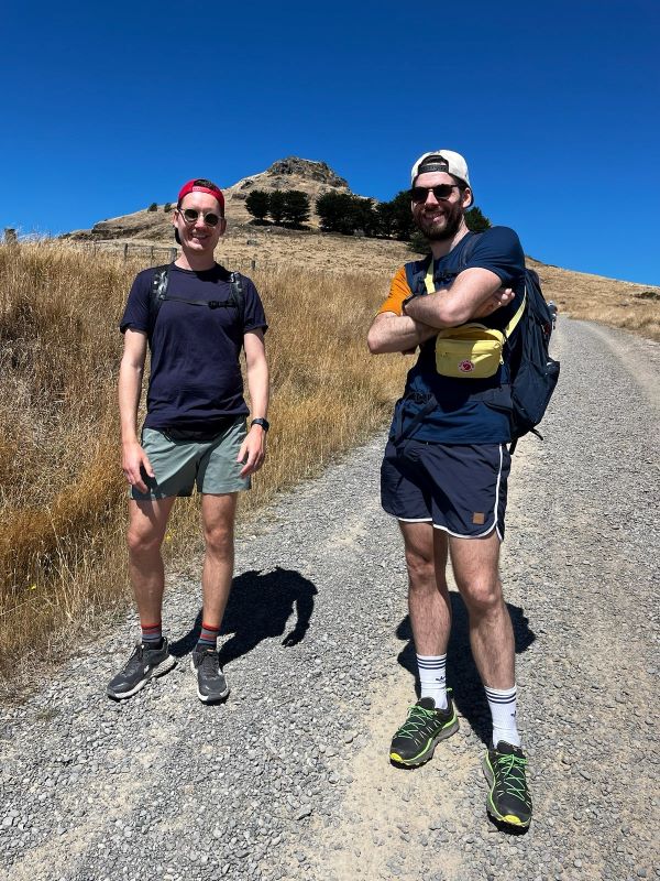 "Manu und ich stehen nebeneinander am Wanderweg und lächeln. Die Landschaft im Hintergrund ist karg und zeigt trockenes Gras und kaum Vegetation."