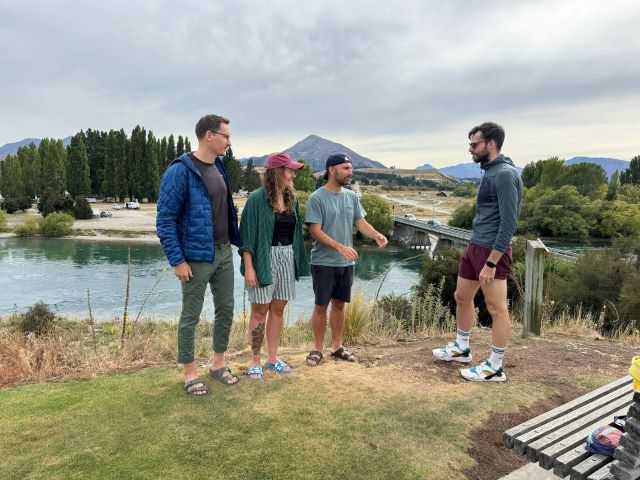 "Vor einem Fluss und einer Berglandschaft steht unsere Gruppe auf der Wiese. Manu, Sophia und Simon stehen am grünen Gras und schauen alle in meine Richtung. Ich stehe auf brauner Erde und sehe grimmig drein."