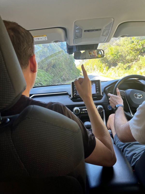 "Foto innerhalb des Autos, aufgenommen vom Rücksatz. Manu am Beifahrersitz hebt den Zeigefinger, ich sitze am Steuer. Im Rückspiegel erkennt man meine Basketballkappe. Die Straße ist umgeben von grünem Wald und die Sonne scheint."