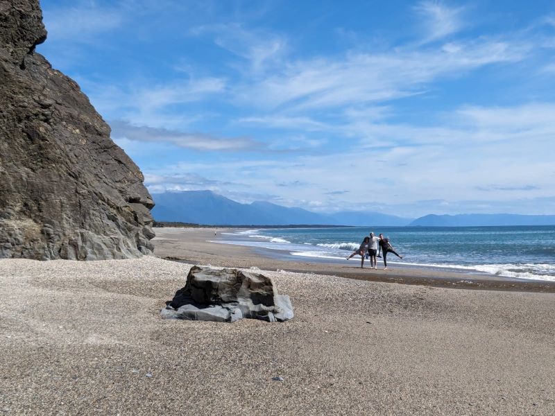 "Der Sandstrand mit dem Ozean und blauen Bergen im Hintergrund unter einem blauen, leicht bewölkten Himmel. AM grauen Strand stehen in der Ferne Sophia, ich und Manu wie wir uns umarmen."