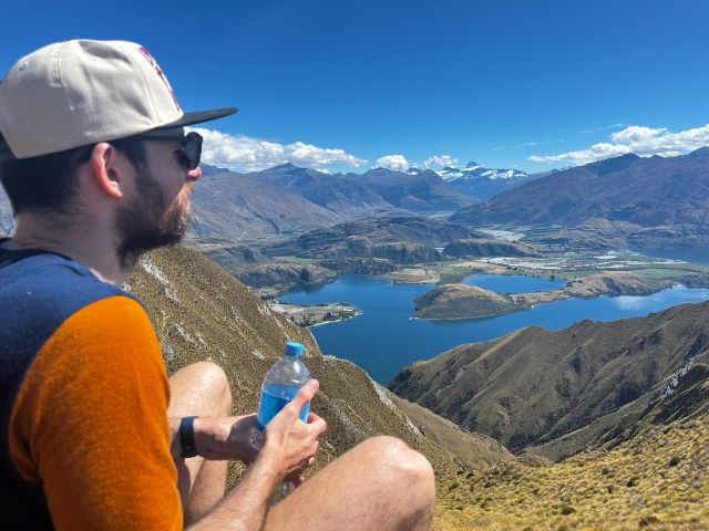 "Ich sitze mit meiner Wasserflasche am Boden und blicke Richtung brauner Berglandschaft. Darunter ist ein blauer See zu erkennen."