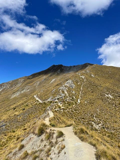 "Vom Roys Peak windet sich ein weiterer Pfad hinauf zu einem weiteren Berggipfel. Der Pfad ist sonnendurchflutet, der Gipfel liegt im Schatten einer Wolke."