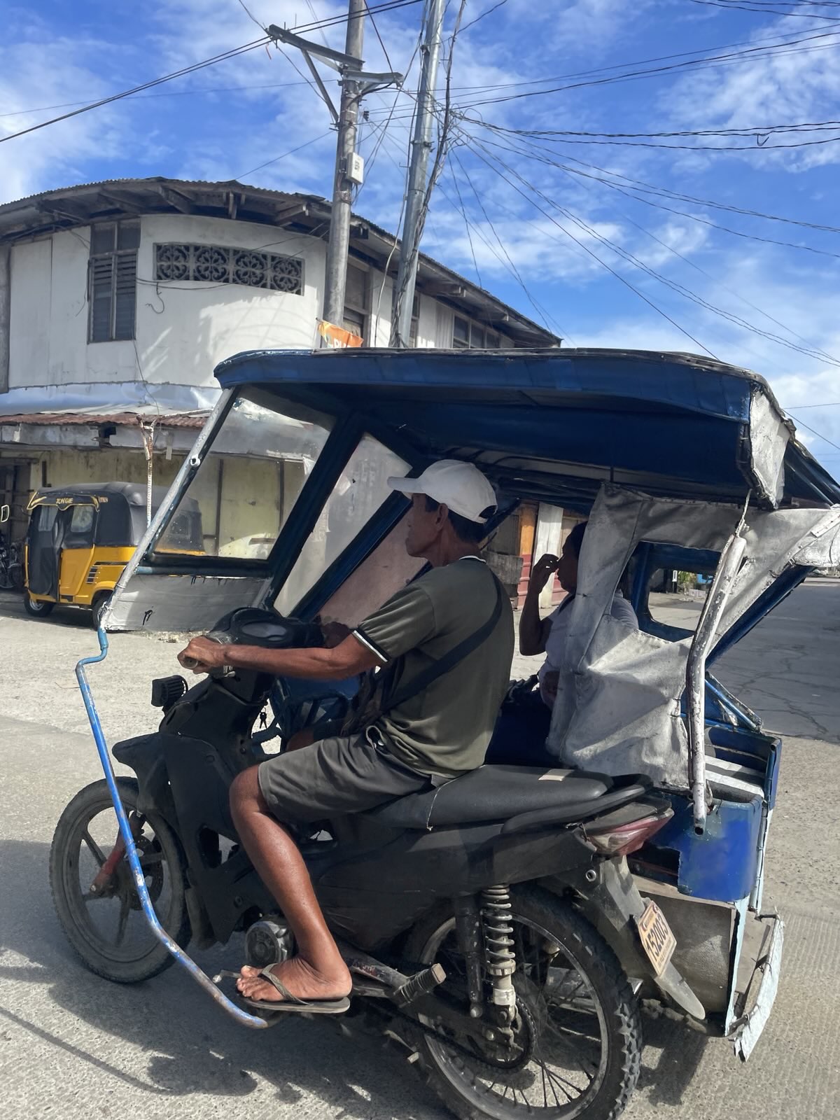 "Seitliche Aufnahme eines Tuktuk-Fahrers, während der Fahrt. Er trägt ein olivfarbenes Poloshirt, eine olivfarbene Hose und eine weiße Kappe. Eine Beifahrerin sitzt neben ihm."