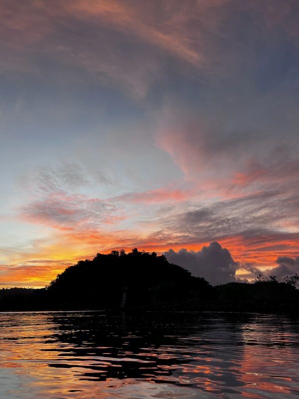 "Das Abendrot über dem Wasser. Einige Wolkenfetzen sind am Himmel zu sehen und wie die Sonne hinter dem Urwald untergeht."