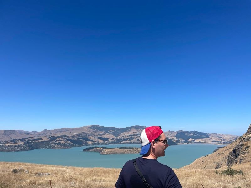 "Manu steht vor einer malerischen Landschaft eines Sees und Bergen im Hintergrund unter einem blauen Himmel. Er trägt eine rot-weiße Kappe mit blauem Schirm"