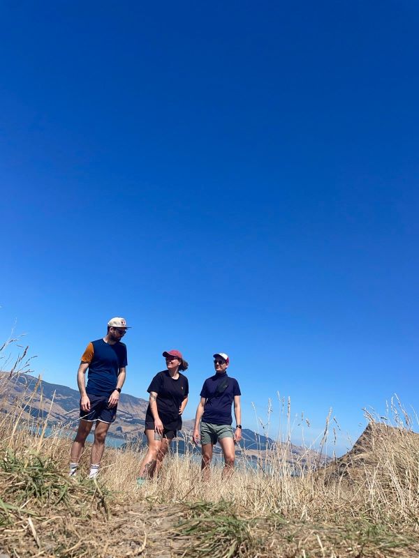 "Ich, Sophia und Manu stehen vor einem Feld mit trockenem Gras unter einem blauen Himmel, mit Bergen und einem blauen See im Hintergrund. Ich trage ein blaues T-Shirt und trage eine Kappe, Sophia trägt ein schwarzes T-Shirt und eine rote Kappe und Manu trägt ein dunkelblaues T-Shirt mit weiß-roter Kappe. Wir sehen einander an."