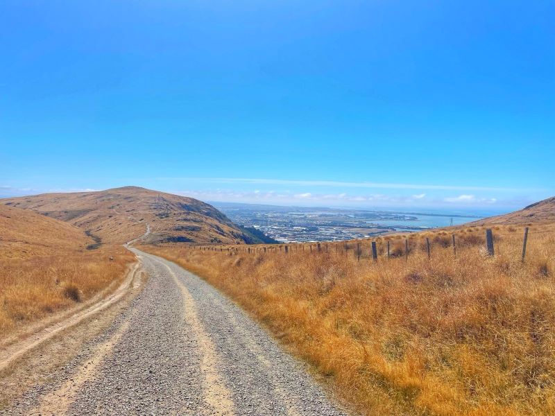 "Eine Schotterstraße die sich durch die trockenen, grasbewachsenen Hügel schlängelt mit einem Blick auf eines kleinen Ortes und der Küste im Hintergrund unter einem blauen Himmel."